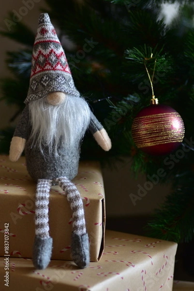 Fototapeta Cozy Christmas scene with a small Nordic gnome sitting on wrapped gifts under a decorated tree. Kraft paper boxes, soft handmade toy and natural holiday decor create a warm festive atmosphere at home.