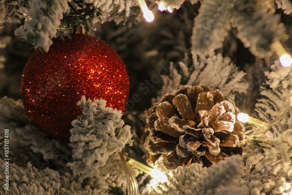 Obraz Festive close-up of a red glitter ornament and a pine cone on a snow-covered Christmas tree with warm holiday lights.