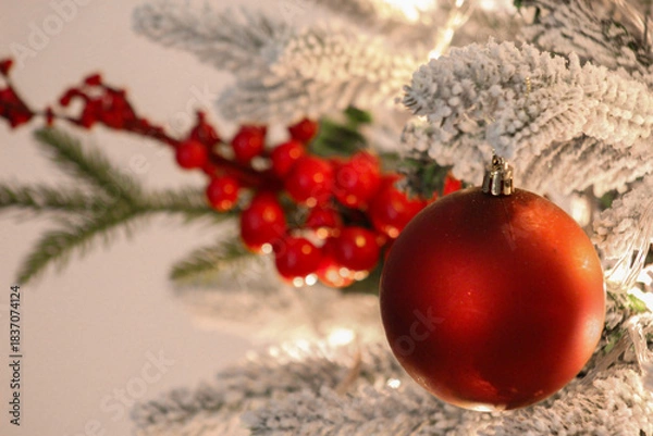 Obraz Close-up of a red Christmas ornament hanging on a snow-covered tree branch with festive berries and warm holiday lights.