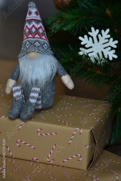 Fototapeta Cozy Christmas scene with a small Nordic gnome sitting on wrapped gifts under a decorated tree. Kraft paper boxes, soft handmade toy and natural holiday decor create a warm festive atmosphere at home.