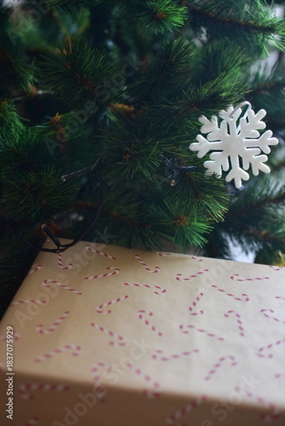Fototapeta White snowflake ornament hanging on green pine branches above a kraft paper gift box with candy cane pattern. Minimal holiday style with natural textures and cozy festive mood.