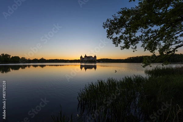 Fototapeta A panoramic, symmetrical shot of Moritzburg Castle at twilight. The sky transitions from a deep blue at the top to an intense orange at the horizon