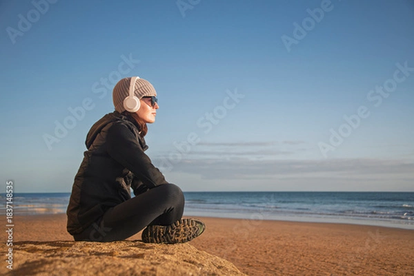 Obraz Woman sitting on a beach wearing headphones and winter jacket, enjoying the ocean view