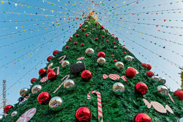 Obraz Giant, brightly decorated outdoor Christmas tree, viewed from below, with red and silver ornaments and festive pennant strings against a blue sky.