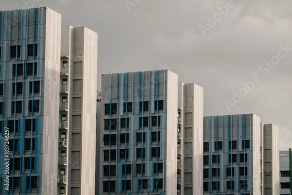 Obraz A row of modern apartment buildings with blue accents under a cloudy sky. The buildings are tall and feature a grid-like facade.