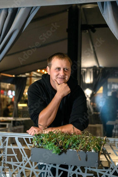 Obraz A thoughtful man in a black hoodie leans on a decorative railing with a planter at night