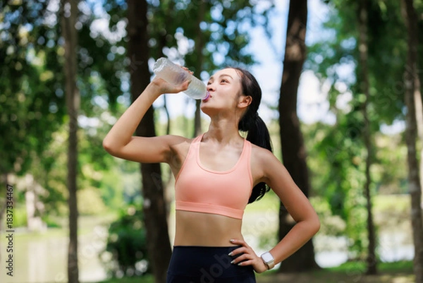 Obraz Young athletic woman drinking water from a bottle, refreshing after an intense workout or jogging session in a lush green park