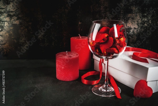 Fototapeta Valentine's day concept, white wrapped gift box with red ribbon, rose flower petals in wine glass, with red candle, on dark stone background, copy space