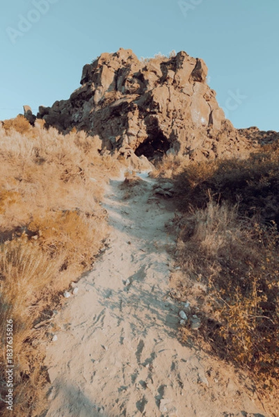 Fototapeta Hiking trail winding through high plains scrub including sagebrush and dappled by late afternoon shade, to large rock formation under a clear blue sky. 