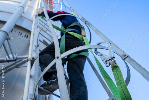Fototapeta A worker or engineer, wearing essential safety equipment including a harness and a yellow hard hat, is captured climbing a fixed vertical ladder on the side of a large industrial silo.