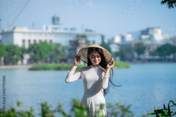 Fototapeta A young Vietnamese woman in a white ao dai and a conical hat standing by Hoan Kiem Lake in Hanoi, Vietnam
