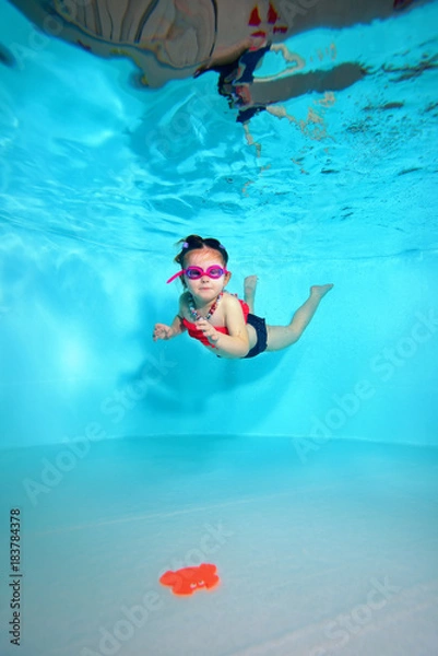 Obraz Child underwater dives to the bottom of the pool after a toy on a blue background. Portrait. Shooting under water. Vertical orientation
