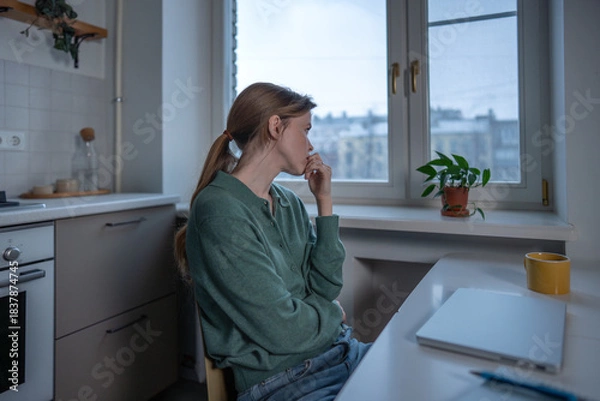 Fototapeta Stressed worried woman sits at table nervously biting nails overwhelmed by thoughts. Tired anxious female pensive looking to window lack motivation, feeling emotional exhaustion, uncertainty at home.