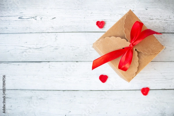Fototapeta Gift box wrapped with a red ribbon placed on a gray background with small hearts beside it, creating a romantic festive composition perfect for Valentine’s Day, love themes and holiday greetings.