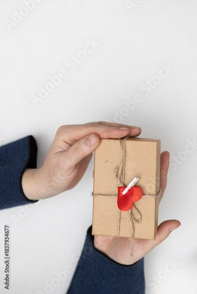 Obraz Two hands are holding a small, wrapped gift. The gift is adorned with twine and a red heart clip. It is perfect for celebrating Saint Valentine's Day.