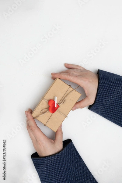 Obraz Two hands hold a small gift box tied with twine and a red heart decoration. The setting is simple and suggests a moment of giving for Valentine's Day.