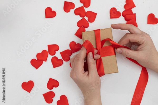 Obraz On a white surface, someone is tying a red ribbon around a small, brown gift box. Many small red hearts are scattered around the box, suggesting a Valentine's Day theme.