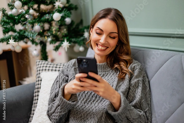 Fototapeta Young happy smiling woman is sitting on couch in her home and using smartphone. Decorated Christmas tree in background.