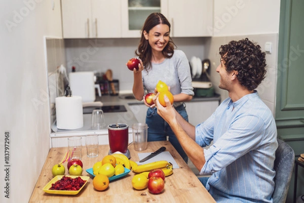 Fototapeta Happy smiling couple share a moment together while preparing homemade fruit drinks, with a countertop full of oranges, apples, and bananas.