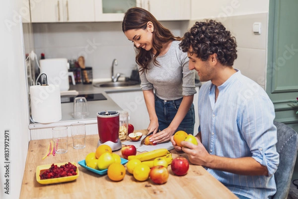 Fototapeta Smiling and relaxed, the two share a moment together while preparing homemade fruit drinks, with a countertop full of oranges, apples, and bananas.