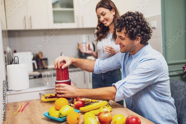 Fototapeta Smiling and relaxed, the two share a moment together while preparing homemade fruit drinks, with a countertop full of oranges, apples, and bananas.