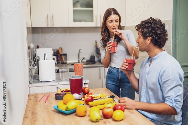 Fototapeta A couple enjoys freshly blended smoothies in their cozy kitchen, surrounded by colorful fruits and a blender filled with a vibrant mix.