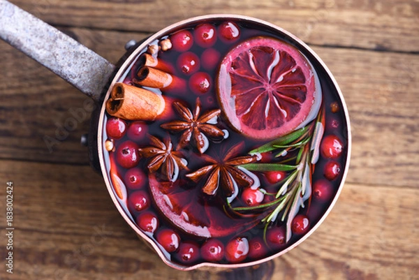 Obraz Copper pot with red mulled wine with slices of orange, cinnamon, star anise, rosemary and cranberries on wooden table. Food photography