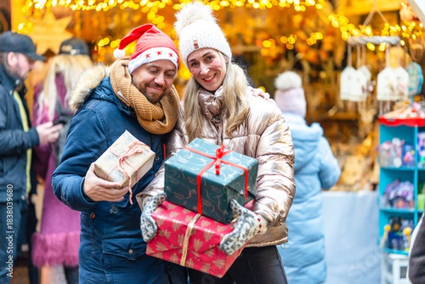 Fototapeta Cheerful couple visiting a Bavarian Christmas market holding presents while enjoying cozy festive winter scenes