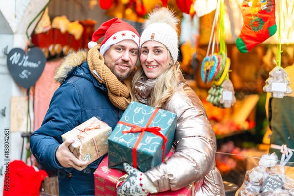 Fototapeta Happy couple enjoying festive lights at a Bavarian Christmas market holding colorful holiday gifts