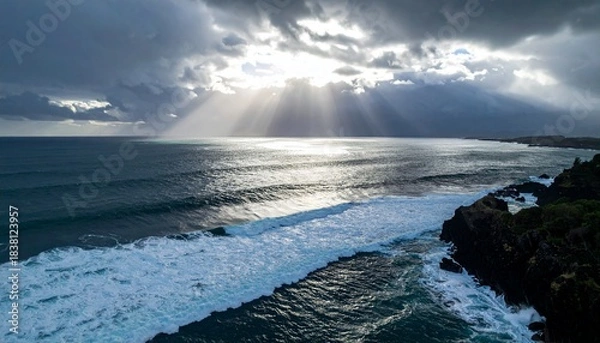 Obraz Dramatic ocean waves crash on a rugged dark rocky coastline under a stormy sky with sun rays breaking through
