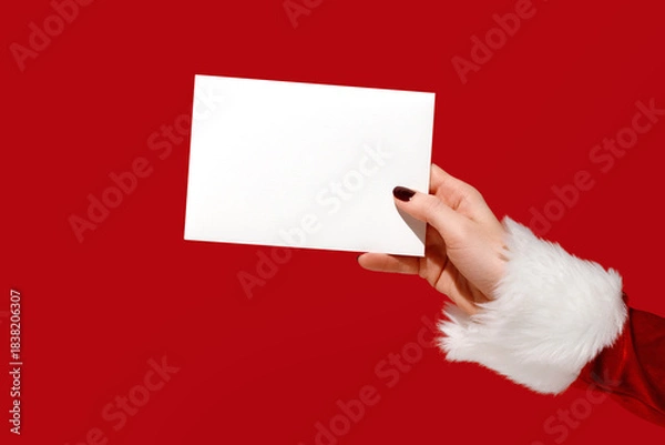 Fototapeta Womans hand in Santa sleeve holding blank card on red background. Christmas mockup