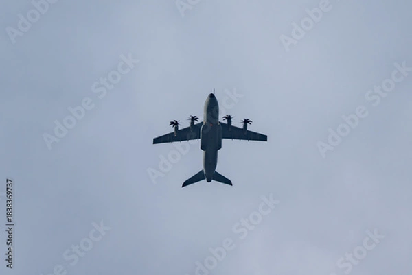 Obraz Four-engine turboprop transport aircraft seen from below against a pale sky. Clean silhouette with wide wings and spinning propellers overhead.