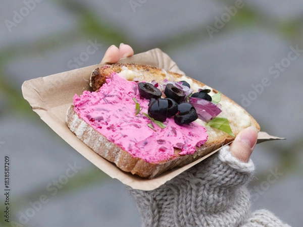 Obraz Hand holding beetroot spread sandwich topped with olives and herbs at Prague Naplavka winter market, showing seasonal cold weather and cozy outdoor food atmosphere.