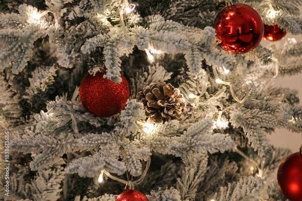 Obraz Snow-covered Christmas tree decorated with red baubles, a pine cone, and warm string lights, creating a festive holiday and winter atmosphere.
