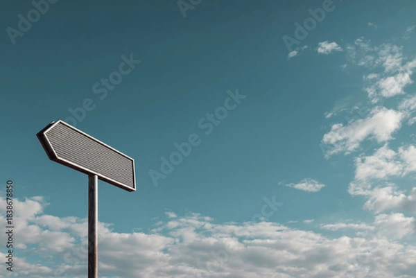 Obraz Textured grey directional arrow sign on a pole, pointing left, against a teal blue sky with white clouds.