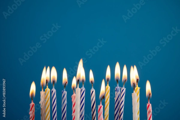 Obraz Lit multicolored birthday candles over blue background creating warm festive glow, with copy space for message or design. Perfect for birthday party invitations or greetings.