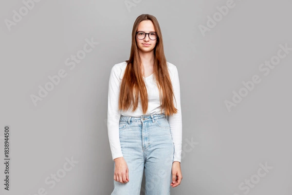 Obraz Smart teenage girl student standing in studio, wearing glasses and casual white clothes, confident young female posing on neutral gray background, education and learning theme, indoor portrait