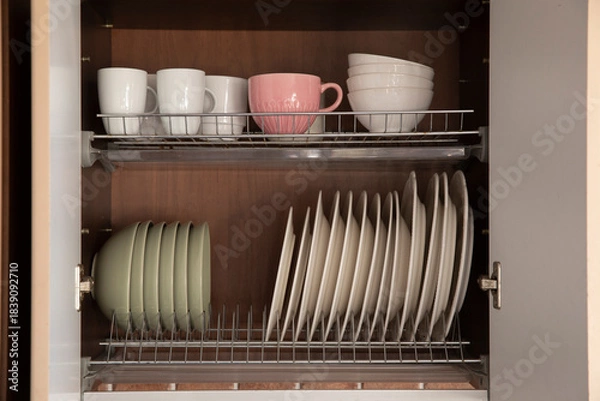 Fototapeta Open kitchen cabinet with neatly arranged plates, bowls and cups on metal drying racks. Organized dish storage, clean minimalist home interior with pastel tableware.