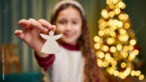 Fototapeta Smiling little girl holding a white angel Christmas ornament in front of glowing holiday tree lights, warm festive atmosphere, shallow depth of field and cozy indoor celebration mood