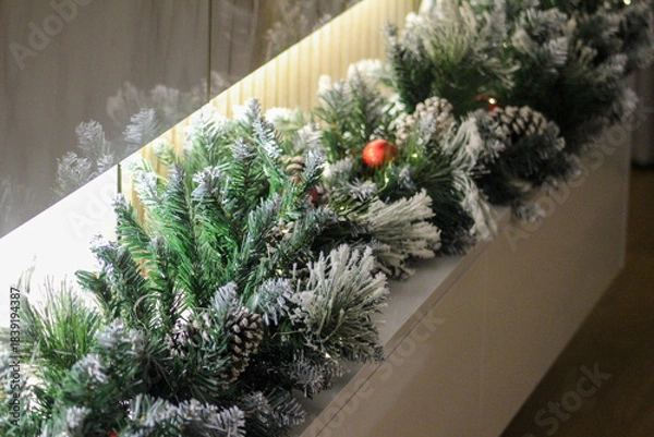Obraz Snow-dusted Christmas garland with pine cones and red ornaments decorates a modern interior, creating a warm and festive holiday atmosphere.