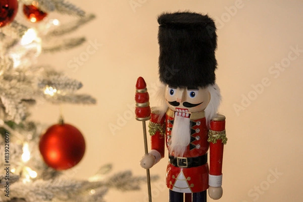 Obraz A decorative nutcracker figure displayed near a Christmas tree with warm white lights and ornaments.