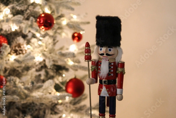 Obraz Wooden nutcracker ornament beside a Christmas tree decorated with red baubles and soft glowing lights.