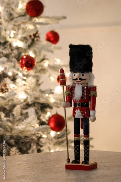 Obraz Wooden nutcracker in red uniform standing beside a softly lit tree decorated with red baubles.