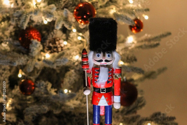 Obraz Festive wooden nutcracker figure standing before a glowing Christmas tree decorated with ornaments and lights.