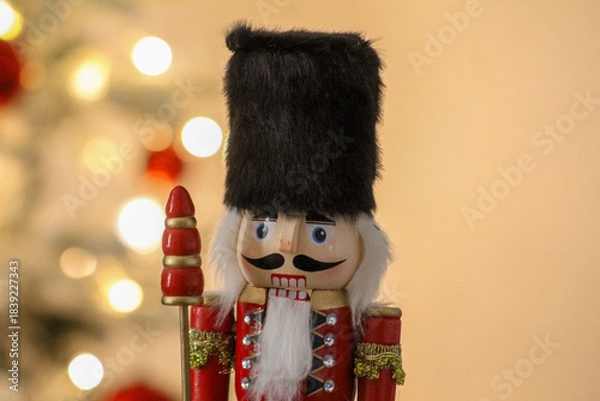 Obraz A wooden nutcracker stands beside a glowing Christmas tree decorated with classic red baubles.