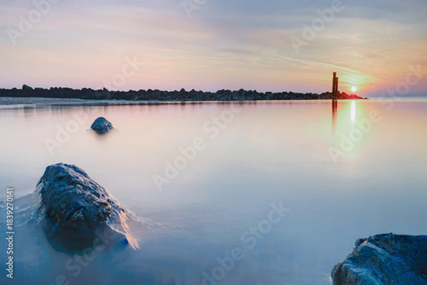 Fototapeta Prominent, textured rock in the shallow foreground, partially submerged in the receding water, reflecting the soft hues of the sky.