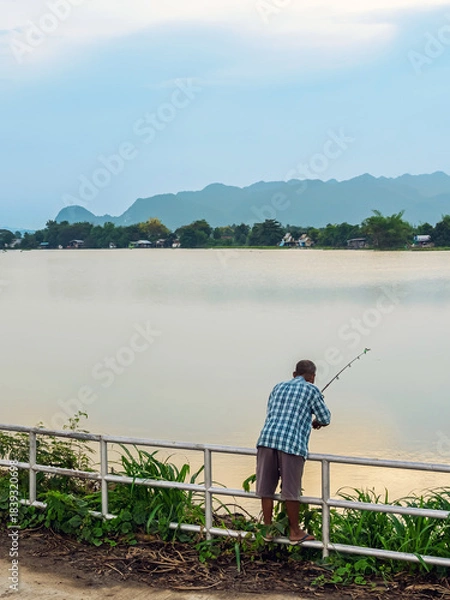 Obraz Elderly fisherman at side of river during sunset and catching fish with spinning. Senior fisherman with fishing rods on footbridge near the lake at summer. Mature man fishing on lake. Adult lifestyle.