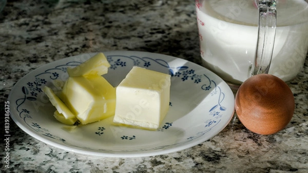 Fototapeta Blocks of butter and a brown egg on a plate with a cup of milk nearby