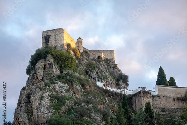 Fototapeta Medieval fortress atop a rugged, rocky cliff under a dramatic, cloudy sky in Olvera, Cadiz, Andalusia, Spain. Below, traditional whitewashed Andalusian houses with terracotta roofs.