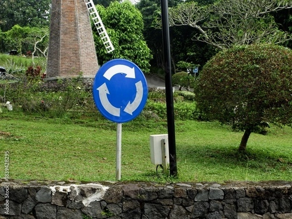 Fototapeta Blue and white circular traffic sign indicating a roundabout or mandatory turn, set in a lush green park with a brick windmill structure in the background. Road sign.